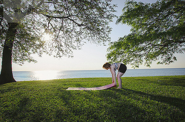 Frau passt Übungsmatte auf Grasfeld im Park gegen Meer und klaren Himmel an