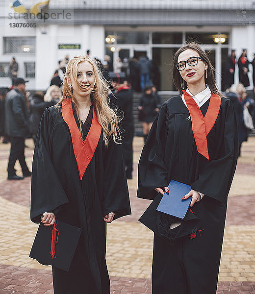 Porträt von glücklichen Studentinnen in Abschlussroben  die gegen das Gebäude stehen