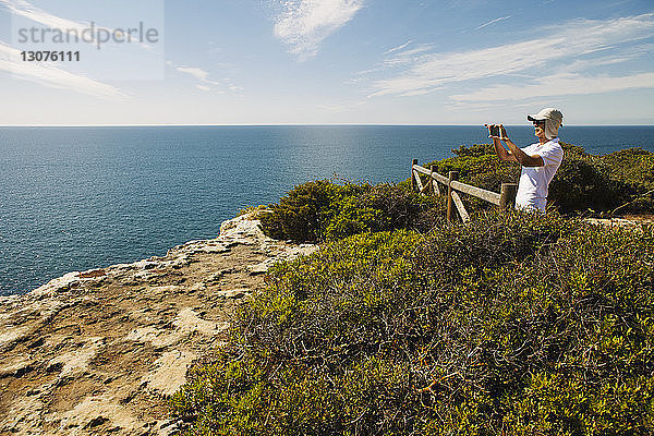 Mann fotografiert Meer durch Telefon  während er auf einer Klippe gegen den Himmel steht