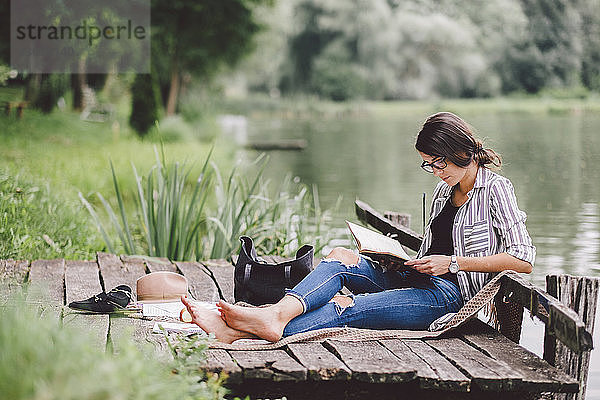 Frau liest Buch  während sie am Pier am See im Wald sitzt