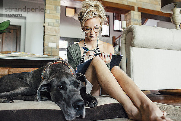 Frau schreibt Tagebuch  während sie bei der Dogge zu Hause auf einem Bett für Haustiere sitzt