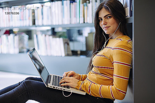 Porträt einer Frau mit Laptop-Computer an Bücherregalen in der Bibliothek sitzend