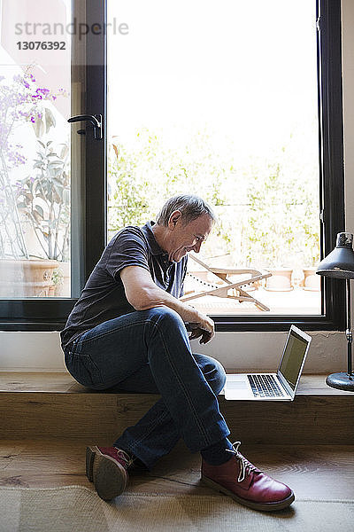 Glücklicher älterer Mann benutzt Laptop  während er zu Hause am Fenster sitzt
