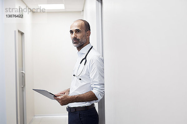 Arzt mit Tablet-Computer schaut weg  während er an der Tür im Korridor steht