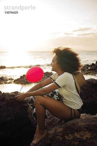 Glückliches Teenager-Mädchen hält Ballon in der Hand  während sie auf felsigem Ufer sitzt