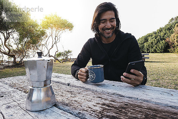 Glücklicher Mann mit Kaffee mit Handy auf Holztisch  während er gegen den Himmel sitzt