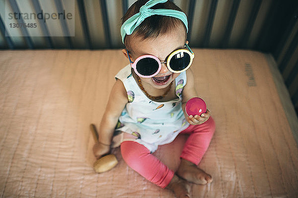 Hochwinkelaufnahme eines verspielten Mädchens mit Sonnenbrille im Kinderbett zu Hause