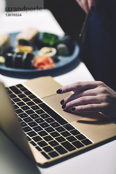 Abgehackte Hand einer Geschäftsfrau  die einen Laptop benutzt  während sie im Heimbüro am Schreibtisch isst