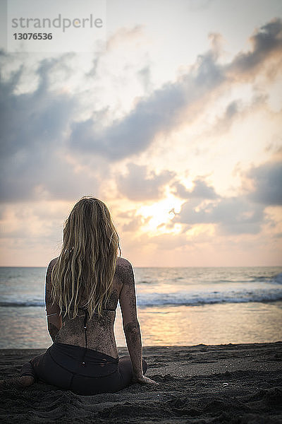 Rückansicht einer am Strand am Strand sitzenden Frau gegen den Himmel bei Sonnenuntergang