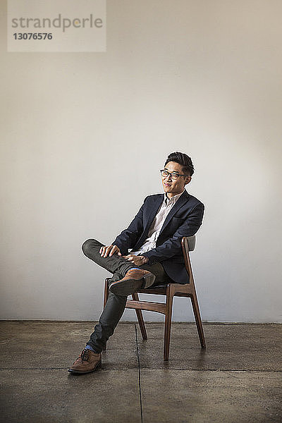 Lächelnder selbstbewusster Geschäftsmann in voller Länge auf Stuhl sitzend an Wand im Büro