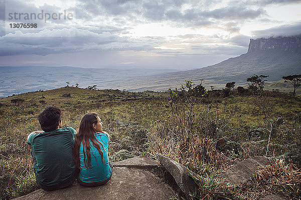 Rückansicht eines Mannes und einer Frau  die auf einem Felsen vor bewölktem Himmel sitzen