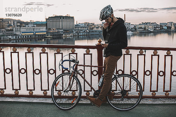 Radfahrer benutzt Smartphone  während er bei Sonnenuntergang mit dem Fahrrad auf der Brücke über den Fluss in der Stadt steht