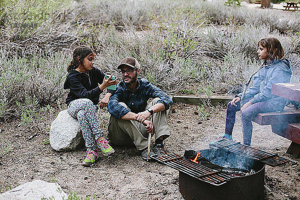 Mädchen sieht Schwester an  die ihrem Vater ein Smartphone zeigt  während sie auf dem Campingplatz sitzt