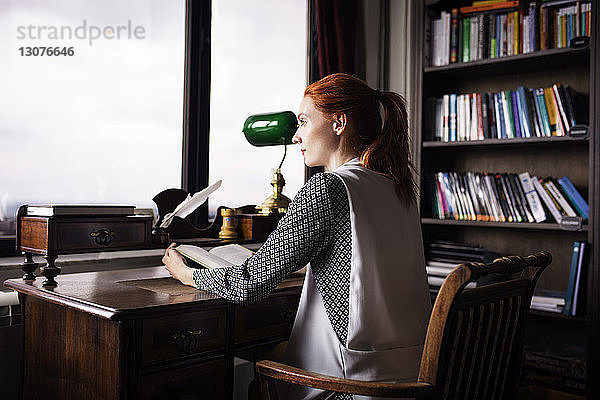 Seitenansicht einer nachdenklichen Geschäftsfrau  die ein Buch in der Hand hält und am Schreibtisch im Kreativbüro sitzt