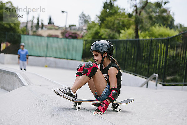 Seitenansicht eines verspielten Mädchens  das an der Sportrampe auf einem Skateboard sitzt