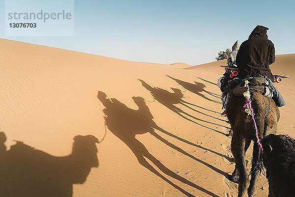 Rückansicht von Freunden  die bei Sonnenschein auf einem Kamel in der Wüste gegen den Himmel sitzen