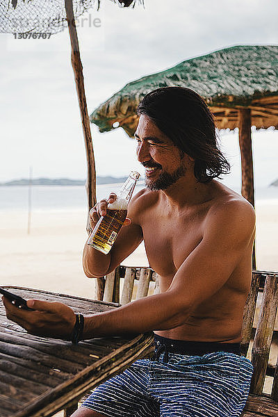 Mann ohne Hemd beim Biertrinken während er am Strand mobil telefoniert