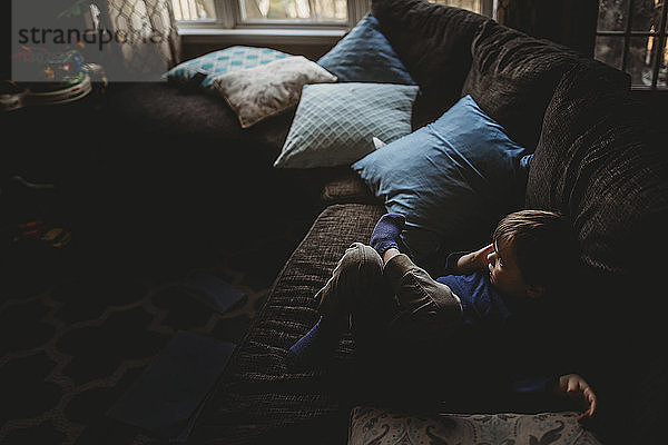 Hochwinkelansicht eines nachdenklichen Jungen  der wegschaut  während er sich zu Hause auf dem Sofa entspannt