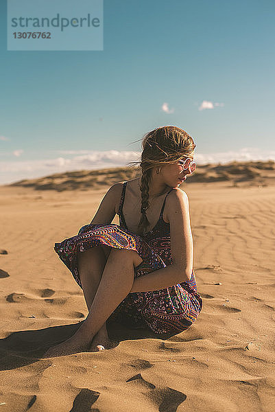 Frau mit Sonnenbrille sitzt am Strand im Sand vor blauem Himmel am sonnigen Tag