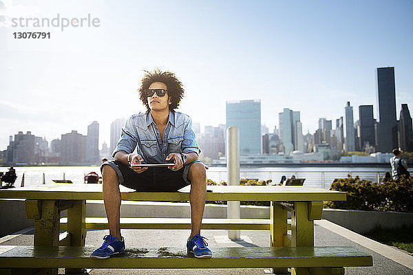 Junger Mann hält Tablette in der Hand  während er auf einer Bank gegen das Stadtbild sitzt