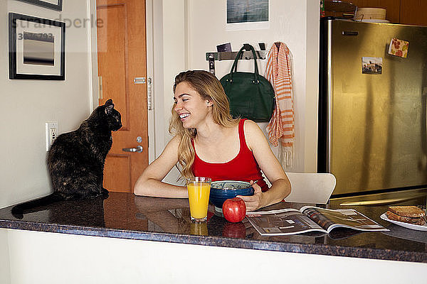 Lächelnde Frau sieht Katze an  während sie beim Frühstück auf der Küchentheke sitzt