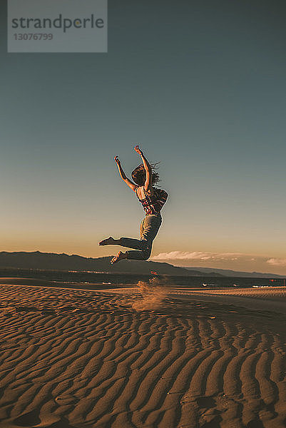 Glückliche Frau springt bei Sonnenuntergang am Strand gegen blauen Himmel