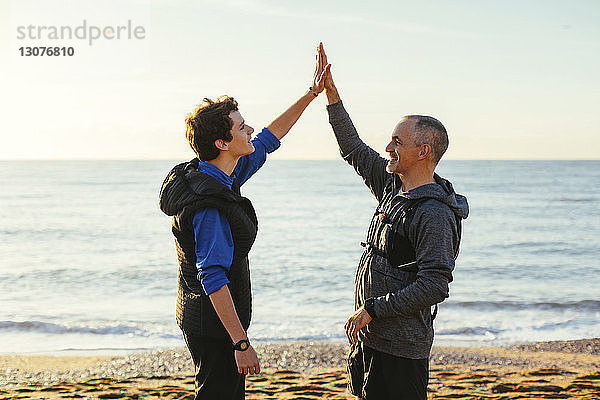 Vater und Sohn machen High-Five  während sie bei Sonnenuntergang am Strand gegen Meer und Himmel stehen