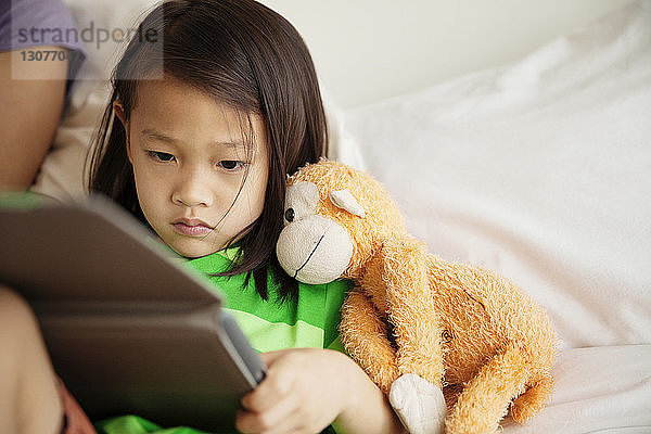 Mädchen mit Plüschtier benutzt Tablet-Computer  während sie neben der Mutter zu Hause im Bett liegt