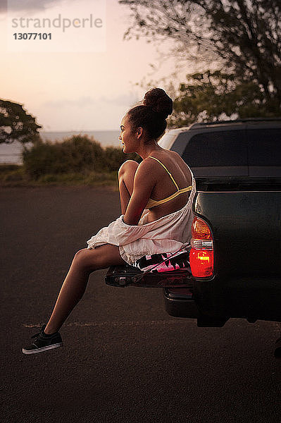 Seitenansicht eines Teenagers  der bei Sonnenuntergang auf einer auf der Straße geparkten LKW-Ladefläche sitzt