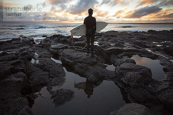 Rückansicht eines Surfers  der ein Surfbrett trägt  während er bei Sonnenuntergang an einer felsigen Küste steht