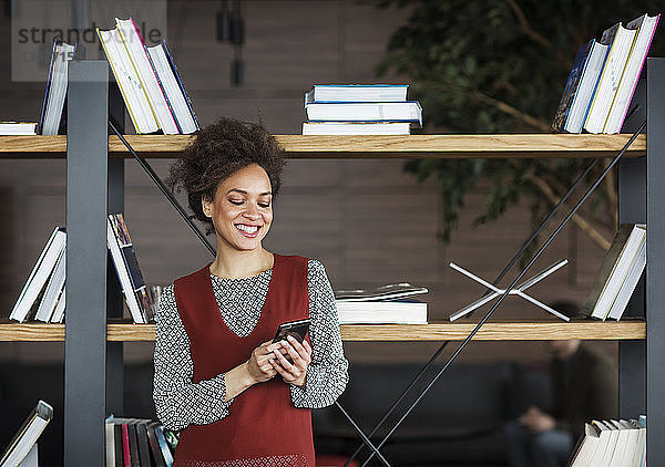 Glückliche Geschäftsfrau benutzt Smartphone  während sie gegen ein Bücherregal steht
