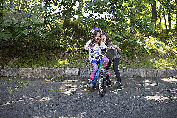 Mädchen hilft Schwester beim Fahrradfahren auf der Straße gegen Bäume im Park