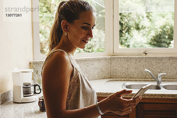 Seitenansicht einer Frau  die ein Smartphone benutzt  während sie in der Küche steht