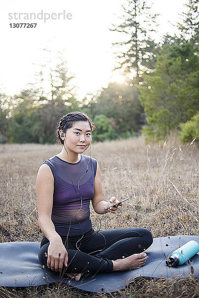 Porträt einer selbstbewussten Frau  die Musik hört  während sie auf einer Übungsmatte auf dem Feld sitzt