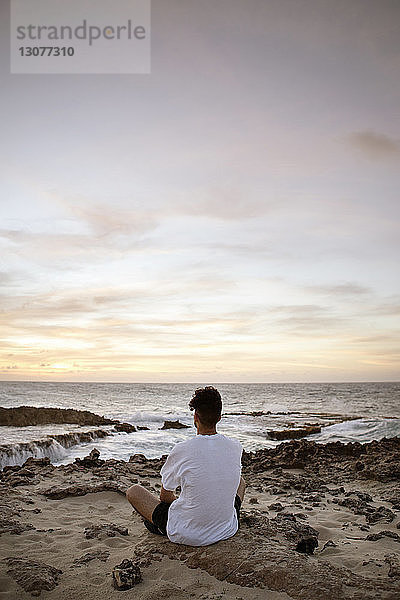 Rückansicht eines Mannes  der auf Sand am Meer sitzt