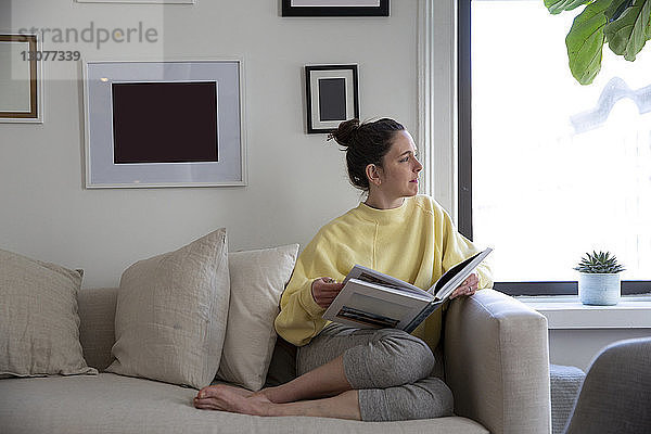Frau hält Zeitschrift in der Hand und schaut weg  während sie zu Hause auf dem Sofa sitzt