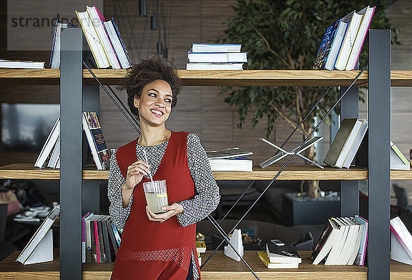 Nachdenkliche glückliche Frau hält Limonade in der Hand  während sie gegen ein Bücherregal steht