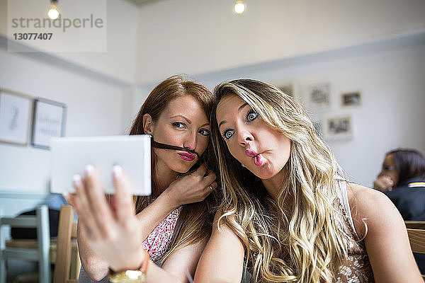 Freundinnen  die Grimassen schneiden  während sie sich im Café per Smartphone unterhalten