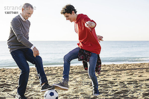 Verspielter Vater und Sohn spielen am Strand bei klarem Himmel Fussball