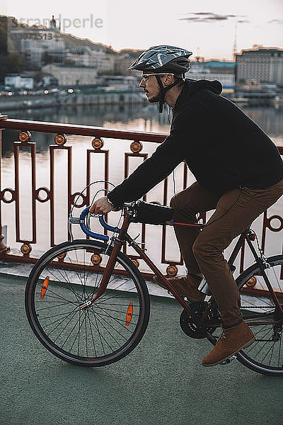 Seitenansicht eines männlichen Radfahrers  der bei Sonnenuntergang auf einer Brücke über einen Fluss in der Stadt Fahrrad fährt
