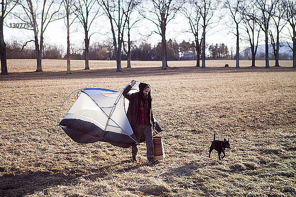 Mann mit tragbarem Zelt in voller Länge beim Spaziergang mit Hund auf dem Feld
