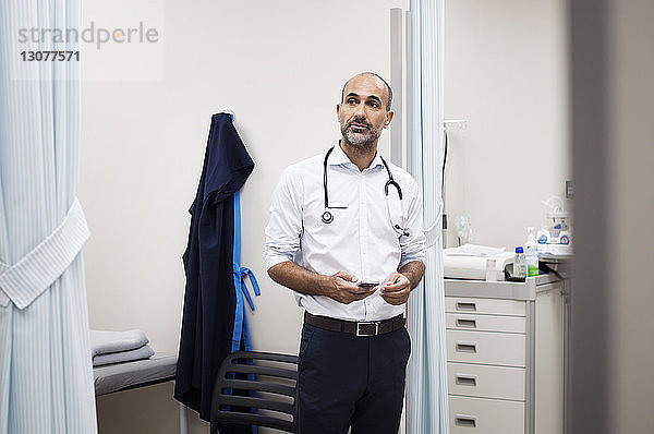 Arzt mit Smartphone schaut weg  während er im Krankenhauszimmer steht