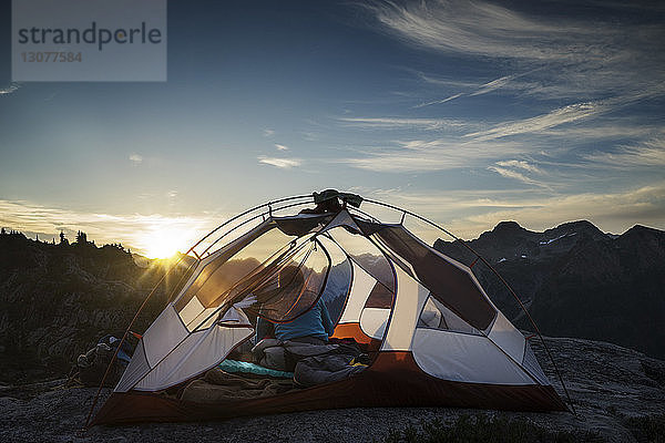 Rückansicht einer Frau  die bei Sonnenuntergang im Zelt vor den Bergen sitzt