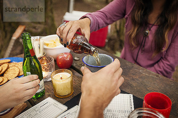 Ausgeschnittenes Bild einer Frau  die Bier für einen Freund ausschenkt  während sie am Picknicktisch im Wald sitzt