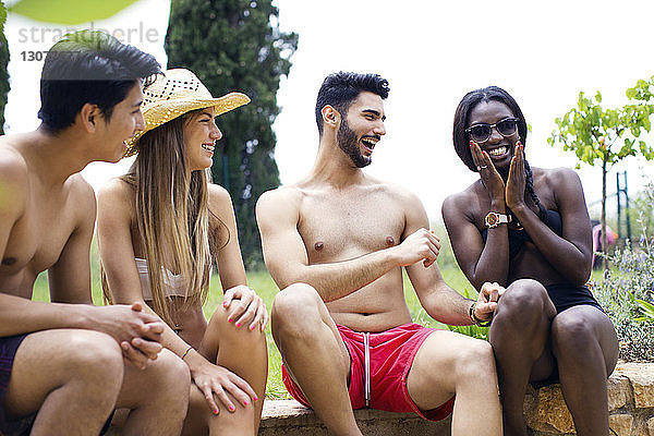 Fröhliche multi-ethnische Freunde in Badebekleidung sitzen auf der Stützmauer im Hof