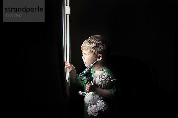 Junge hält Spielzeug in der Hand  während er zu Hause in der Dunkelkammer durchs Fenster schaut