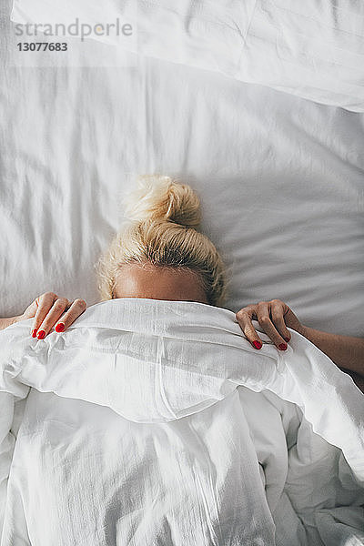 Draufsicht einer Frau  die ihr Gesicht mit einer Decke auf dem Bett bedeckt