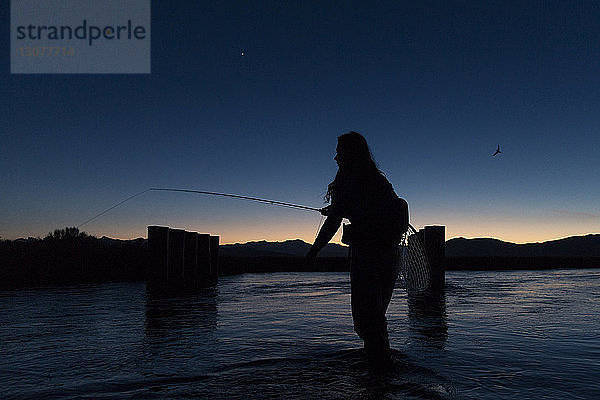 Silhouette einer jungen Frau beim Fliegenfischen  die in der Dämmerung im Fluss Owens gegen den Himmel steht