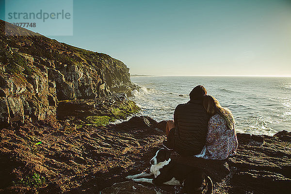 Ehepaar und Hund sitzen auf einem Felsen am Meer vor klarem Himmel