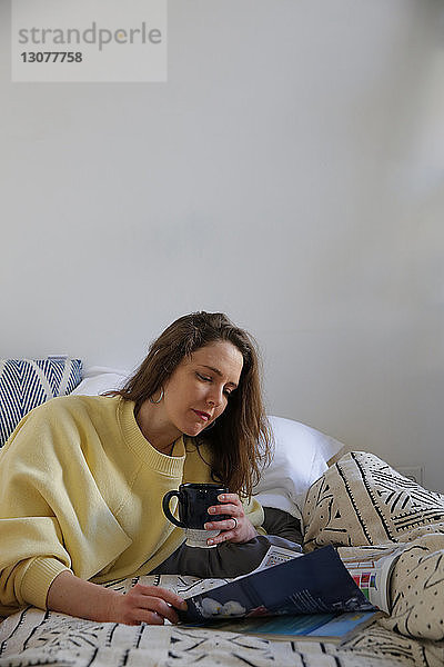 Frau hält Kaffeetasse  während sie zu Hause auf dem Bett eine Zeitschrift liest